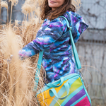 Person wearing a colorful jacket with a multicolored bag, standing among tall dried plants.