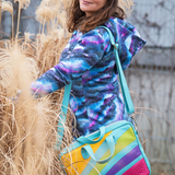 Person wearing a colorful jacket with a multicolored bag, standing among tall dried plants.