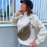 A person wearing a beige knit sweater and blue jeans is fashionably accessorized with a brown leather bum bag (fanny pack) with a zipper closure. The person has a hat on, and the image captures them in a casual, outdoor setting.
