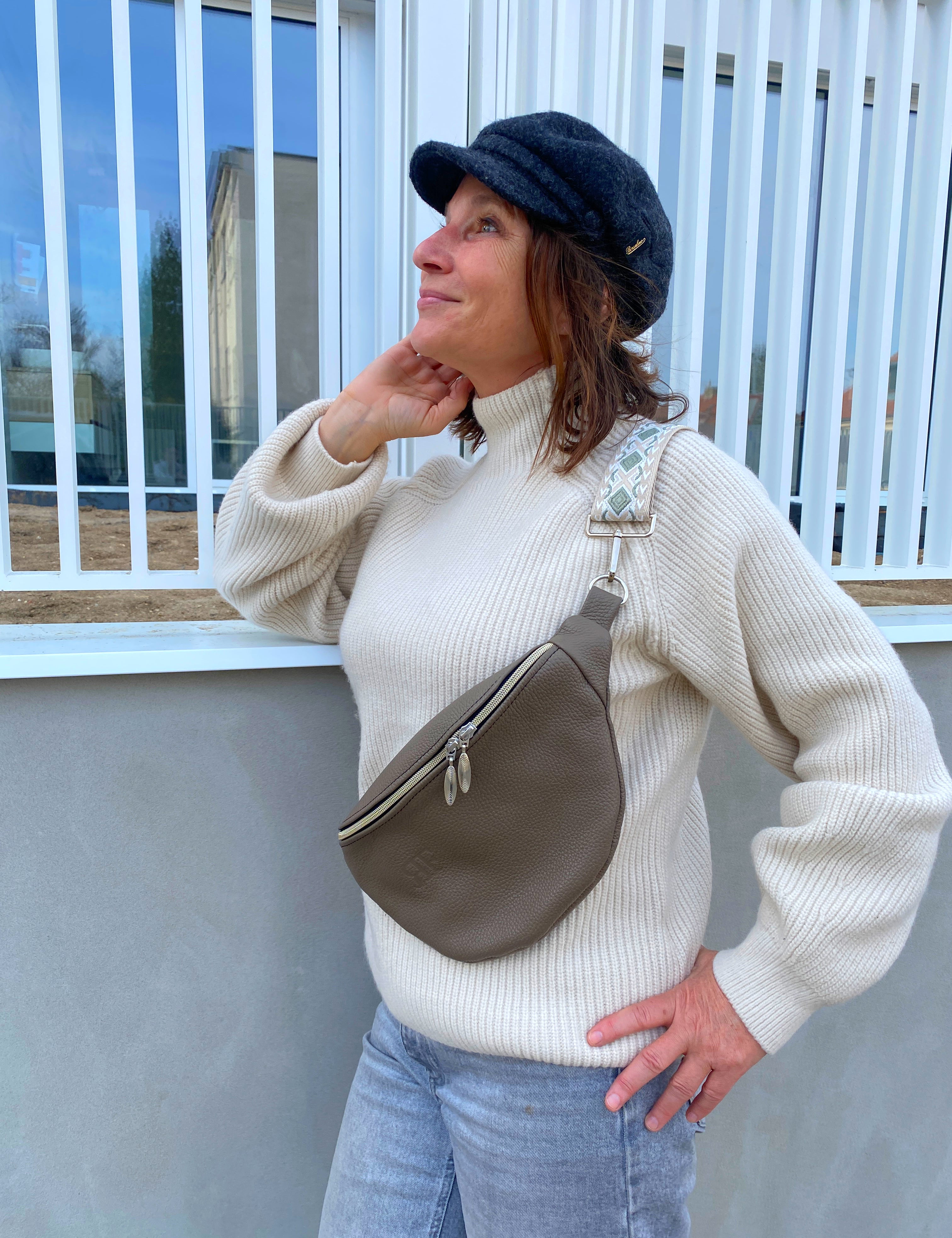 A person wearing a beige knit sweater and blue jeans is fashionably accessorized with a brown leather bum bag (fanny pack) with a zipper closure. The person has a hat on, and the image captures them in a casual, outdoor setting.