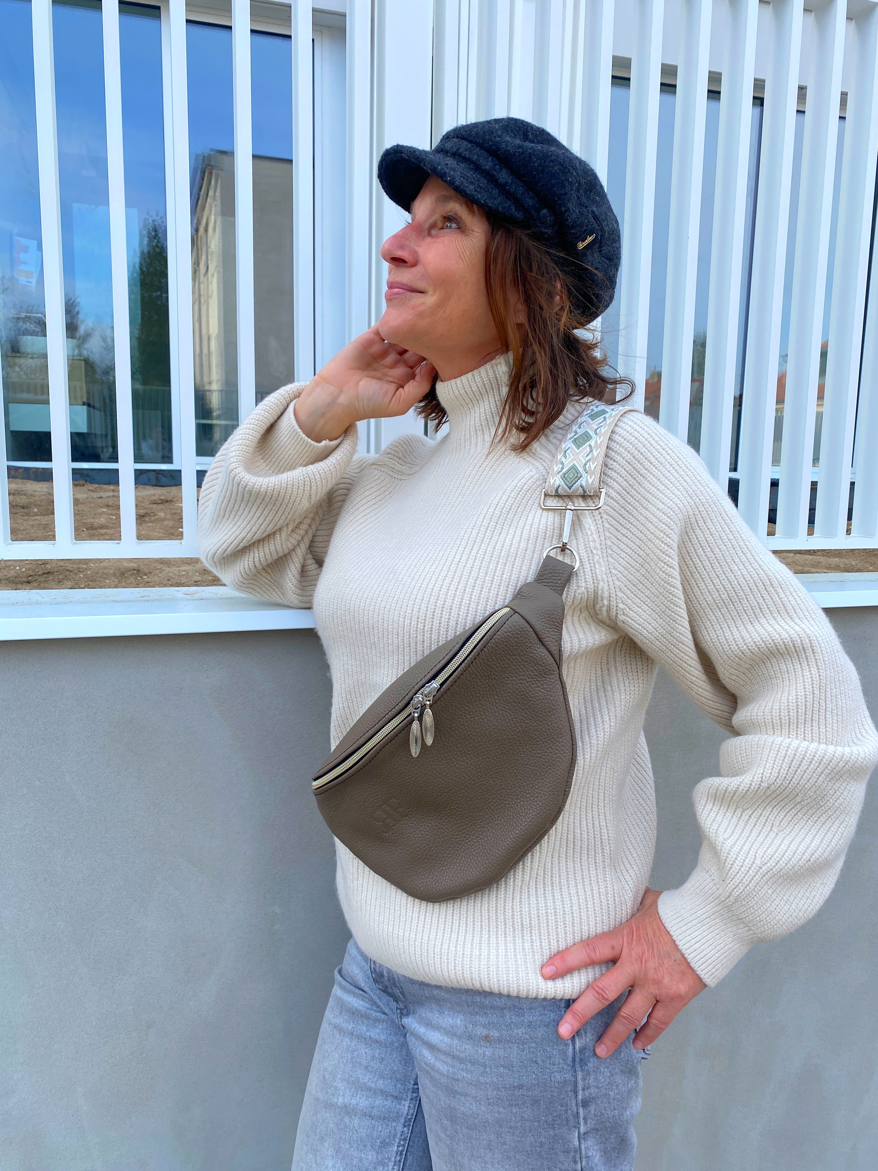 A person wearing a beige knit sweater and blue jeans is fashionably accessorized with a brown leather bum bag (fanny pack) with a zipper closure. The person has a hat on, and the image captures them in a casual, outdoor setting.