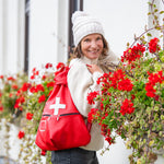 Woman holding a red bag with a white cross, standing in front of a building with flowers.