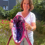 Woman holding a pink and purple patterned bag outdoors with greenery and flowers in the background