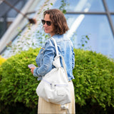 A woman in a light blue denim jacket and beige skirt stands outdoors in front of green bushes and a modern glass building, carrying a large white shoulder bag.