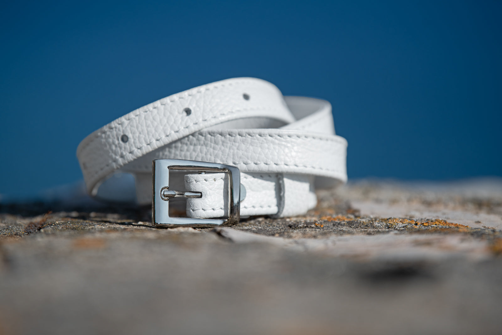 White leather belt with a silver buckle on a textured surface with a blue background