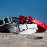 Three dleather belts in black, white, and red on a textured surface with a blue sky background.