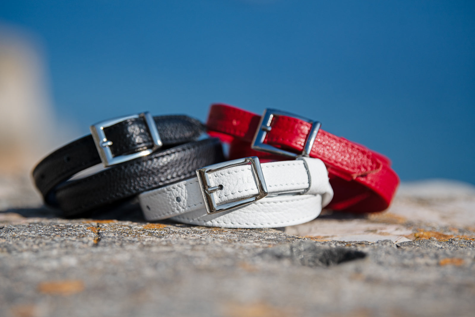 Three dleather belts in black, white, and red on a textured surface with a blue sky background.