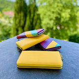 Yellow wallet, pink and purple pen, and pink and yellow wallet on a blue surface with greenery in the background