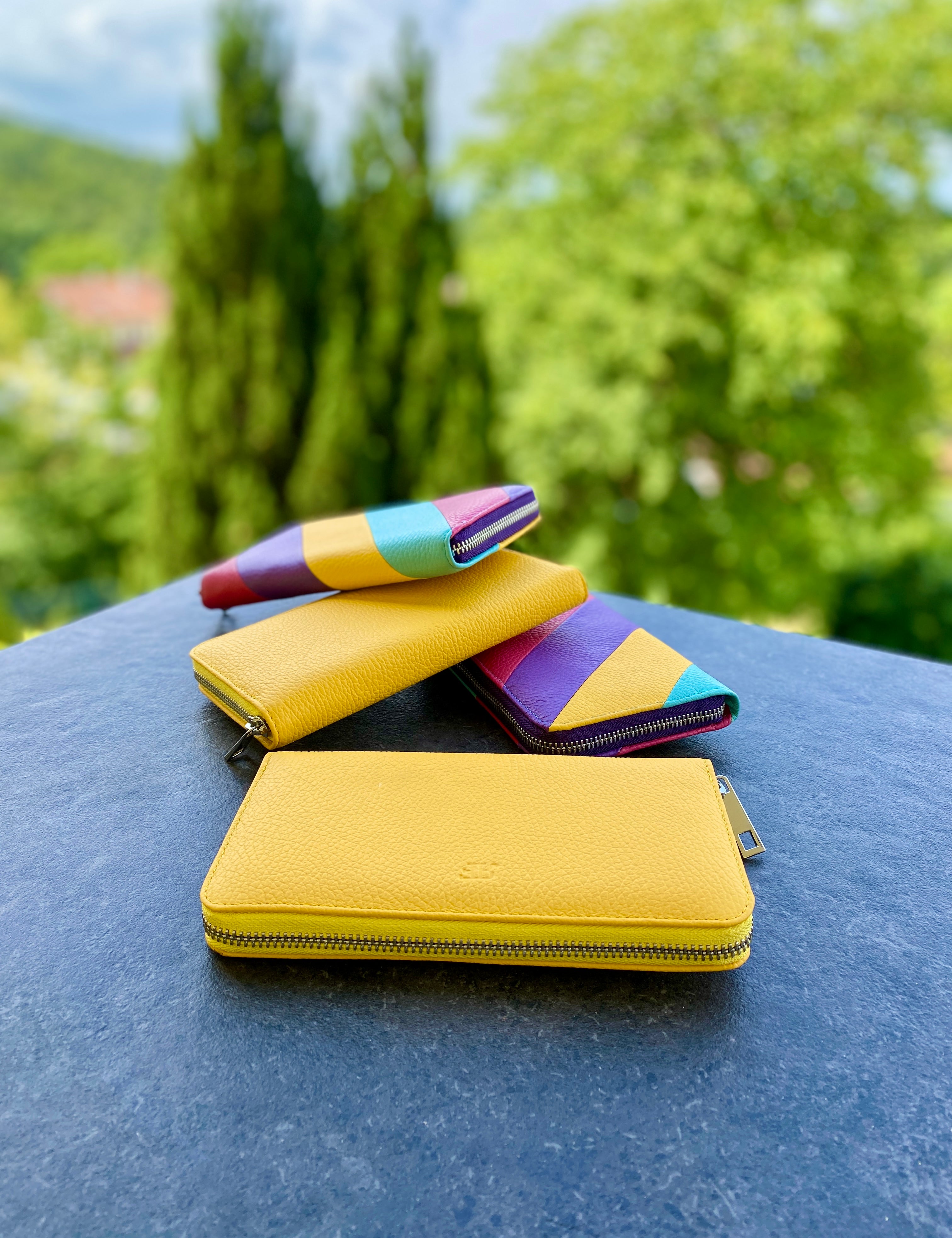 Yellow wallet, pink and purple pen, and pink and yellow wallet on a blue surface with greenery in the background