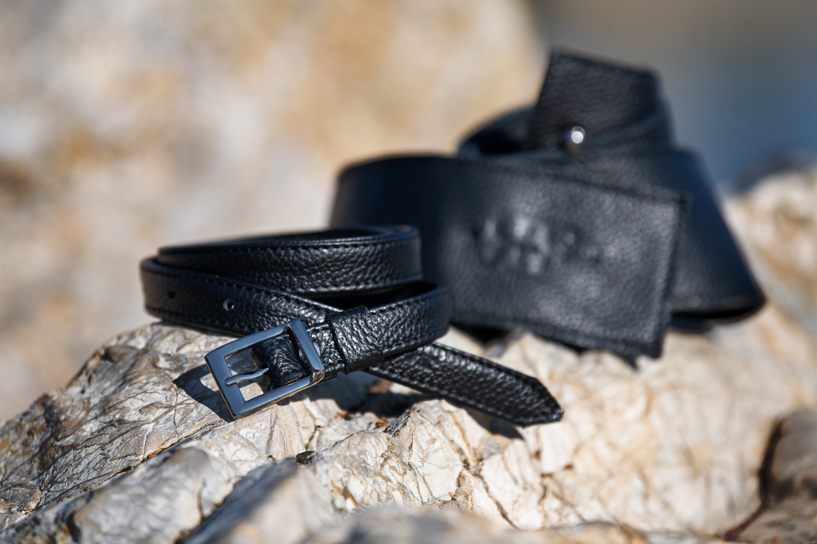 Two black leather belts on a textured stone surface with a blurred natural background.